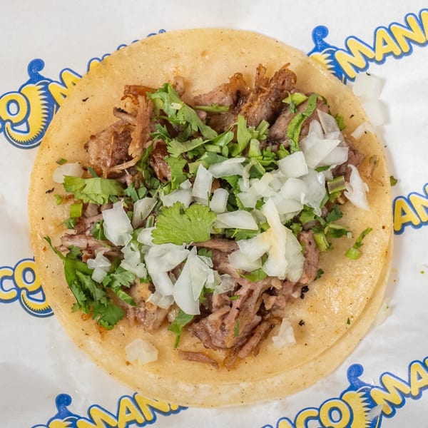 A TacoMania taco with shredded meat, chopped onions, and cilantro on a corn tortilla.