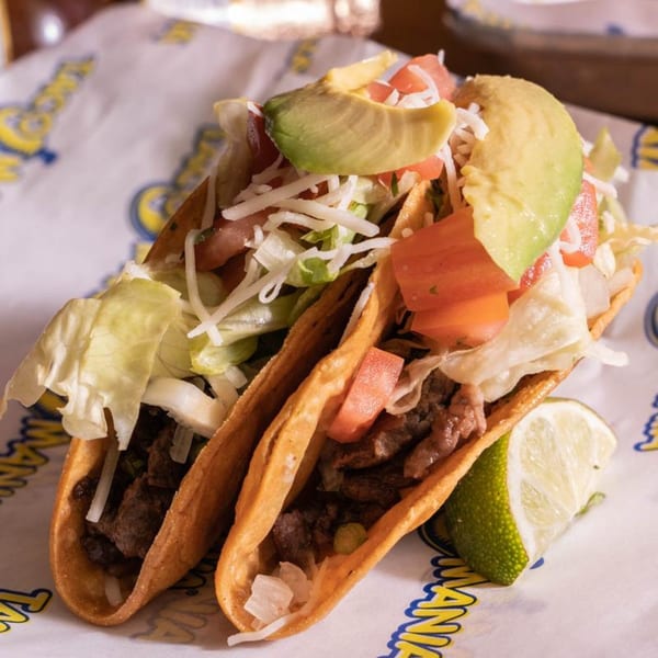 Two TacoMania tacos with beef, lettuce, cheese, tomatoes, avocado, and a lime wedge.