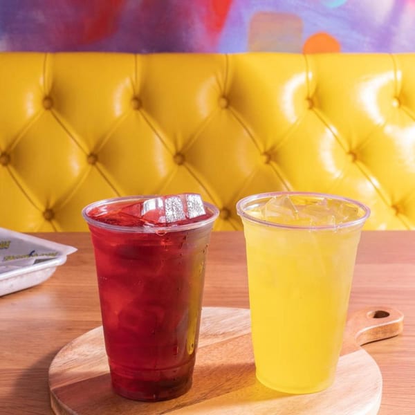 Two iced drinks, one red and one yellow, on a wooden board with a yellow booth in the background.
