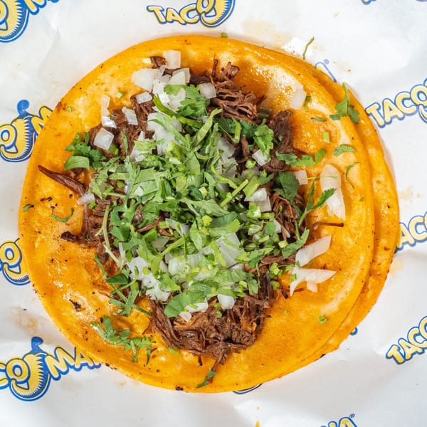 A TacoMania taco with shredded beef, diced onions, and cilantro on a yellow tortilla.