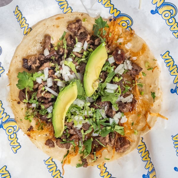 TacoMania carne asada taco with avocado, onions, and cilantro on branded paper.