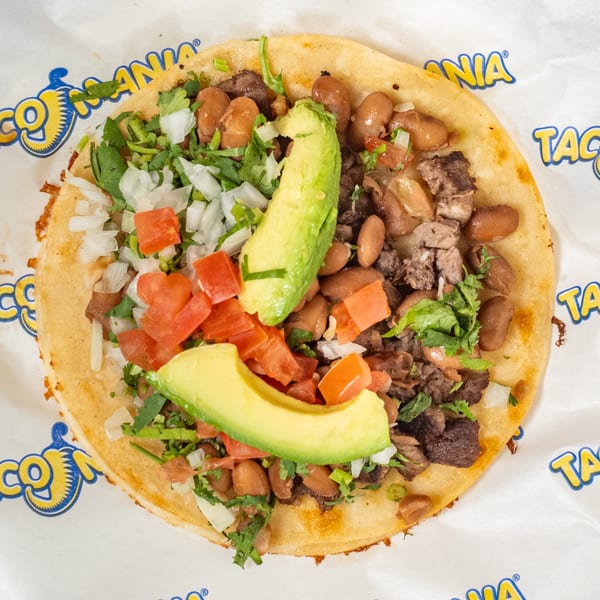 TacoMania taco with meat, beans, diced tomatoes, onions, cilantro, and avocado slices on branded paper.