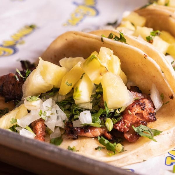 Three TacoMania tacos filled with meat, cilantro, diced onions, and pineapple chunks on a tray.