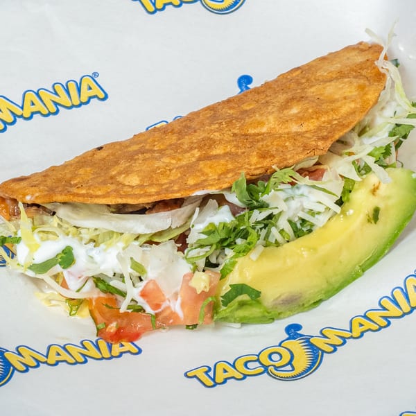 A crispy TacoMania taco filled with meat, lettuce, tomatoes, sour cream, and a slice of avocado.