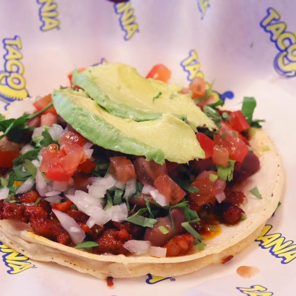 A TacoMania taco filled with meat, diced tomatoes, onions, cilantro, and sliced avocado.